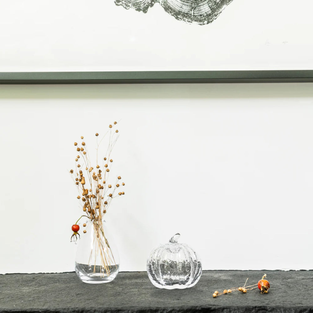 A clear glass pumpkin-shaped vase and a clear glass bud vase with dried botanical stems and a small red berry, arranged on a dark tabletop against a white wall.