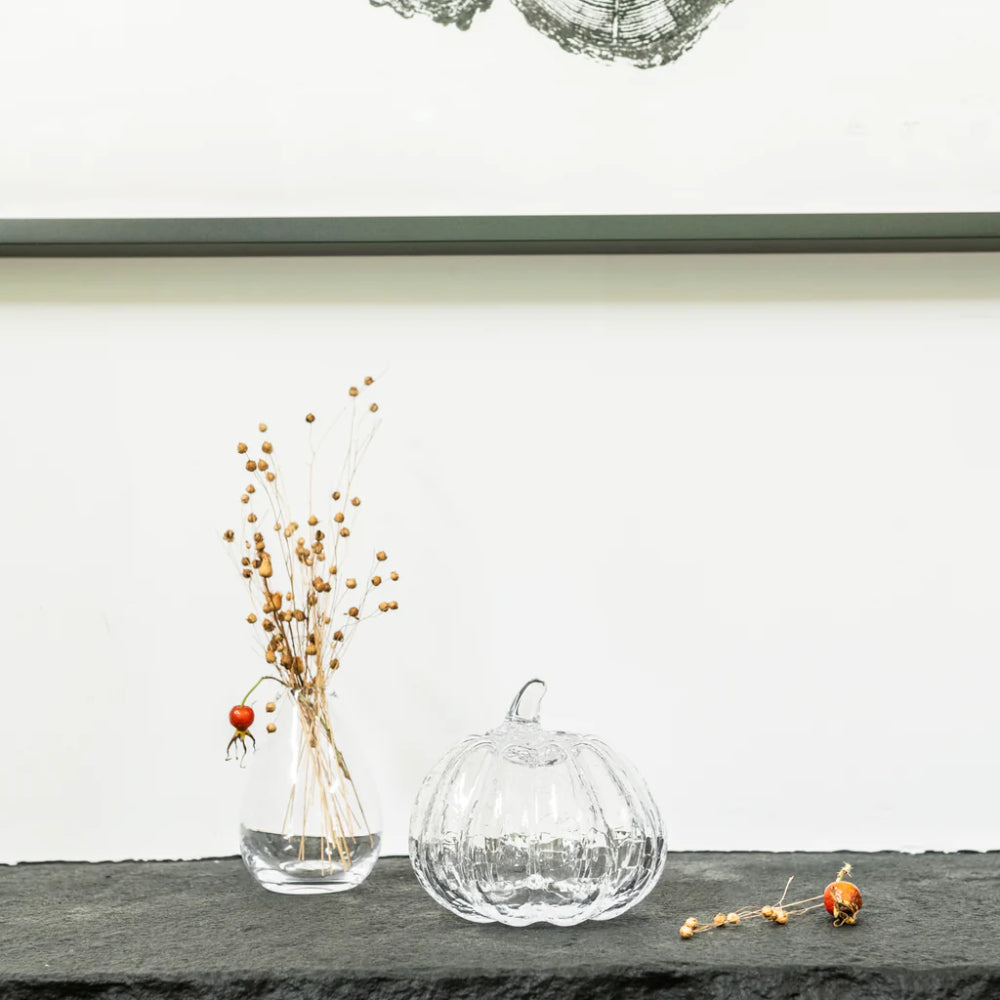 A clear ribbed glass pumpkin vase and a matching clear glass bud vase with dried botanical stems, styled on a dark slate surface against a white wall.