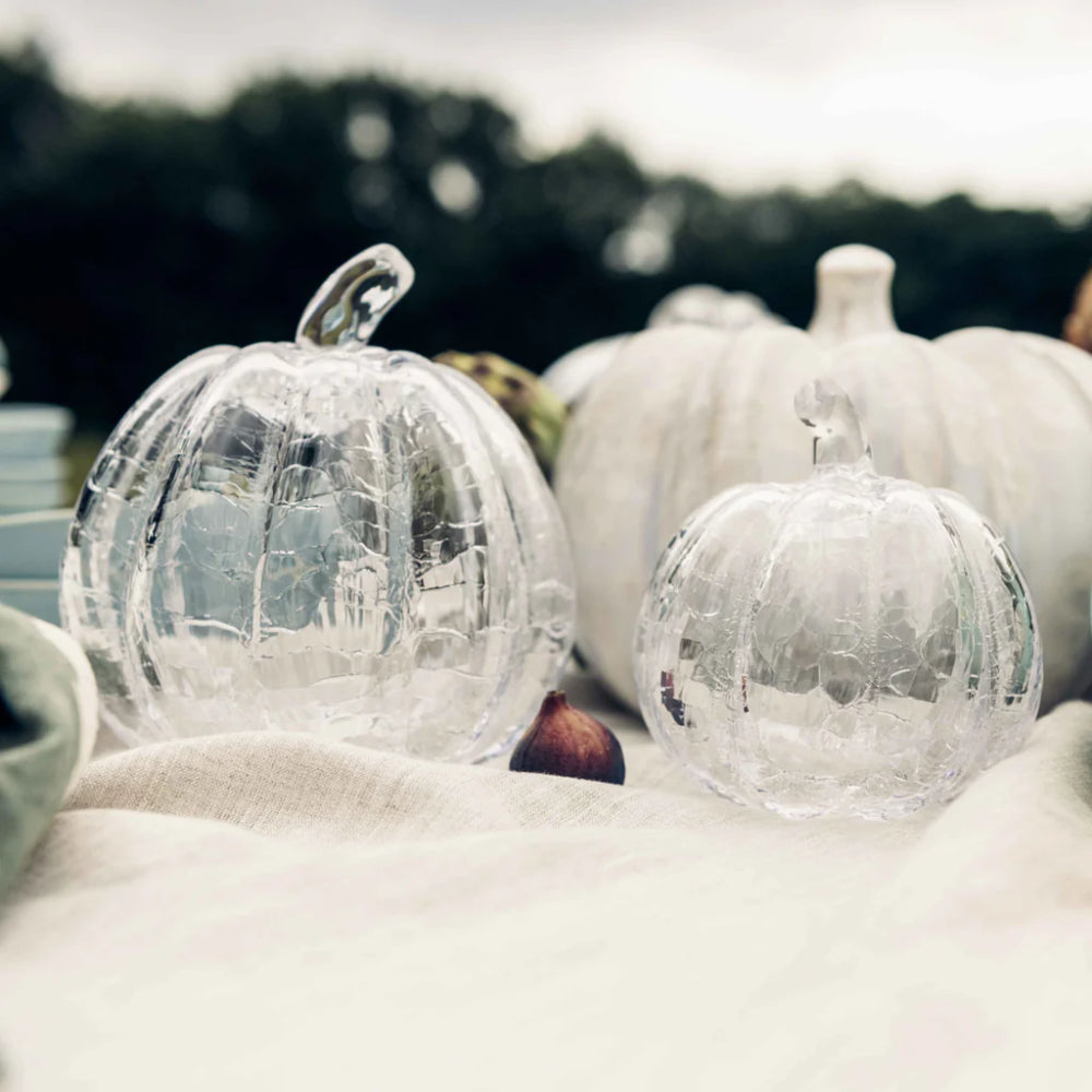 Two clear crackle-glass pumpkins with stems, one large and one small, displayed on a white fabric surface outdoors with blurred white pumpkins and trees in the background.