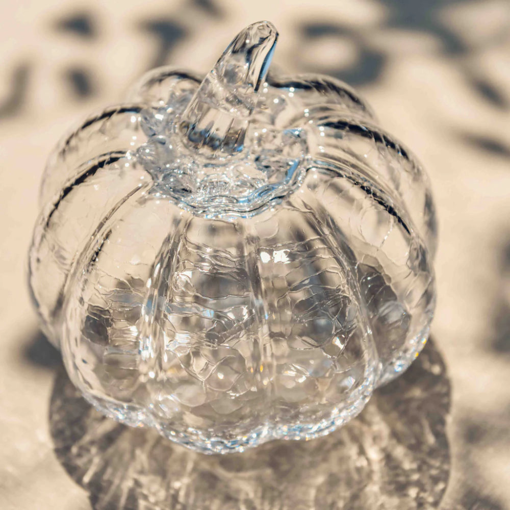 A clear glass pumpkin-shaped decorative container with a textured, hammered surface and a short stem, photographed on a light beige surface with soft shadows.