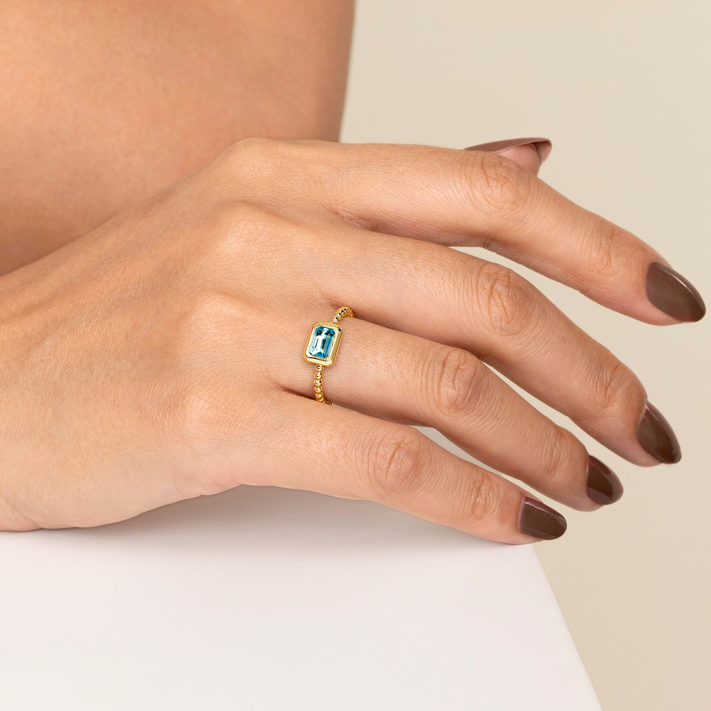 Hand wearing a gold ring with a blue gemstone on a neutral background