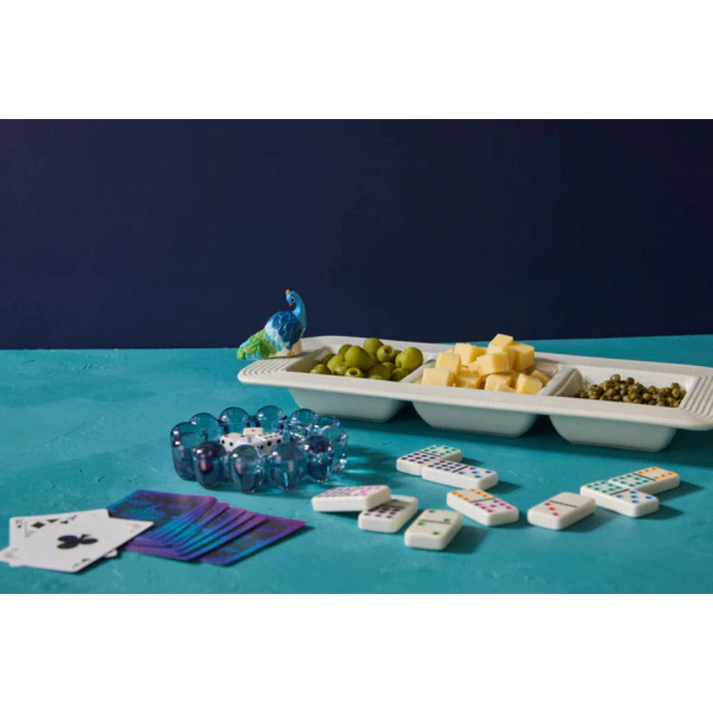 Colorful peacock figurine mini  on a on a white 3 tier serving tray with cheese and all in them and game pieces on a blue table 