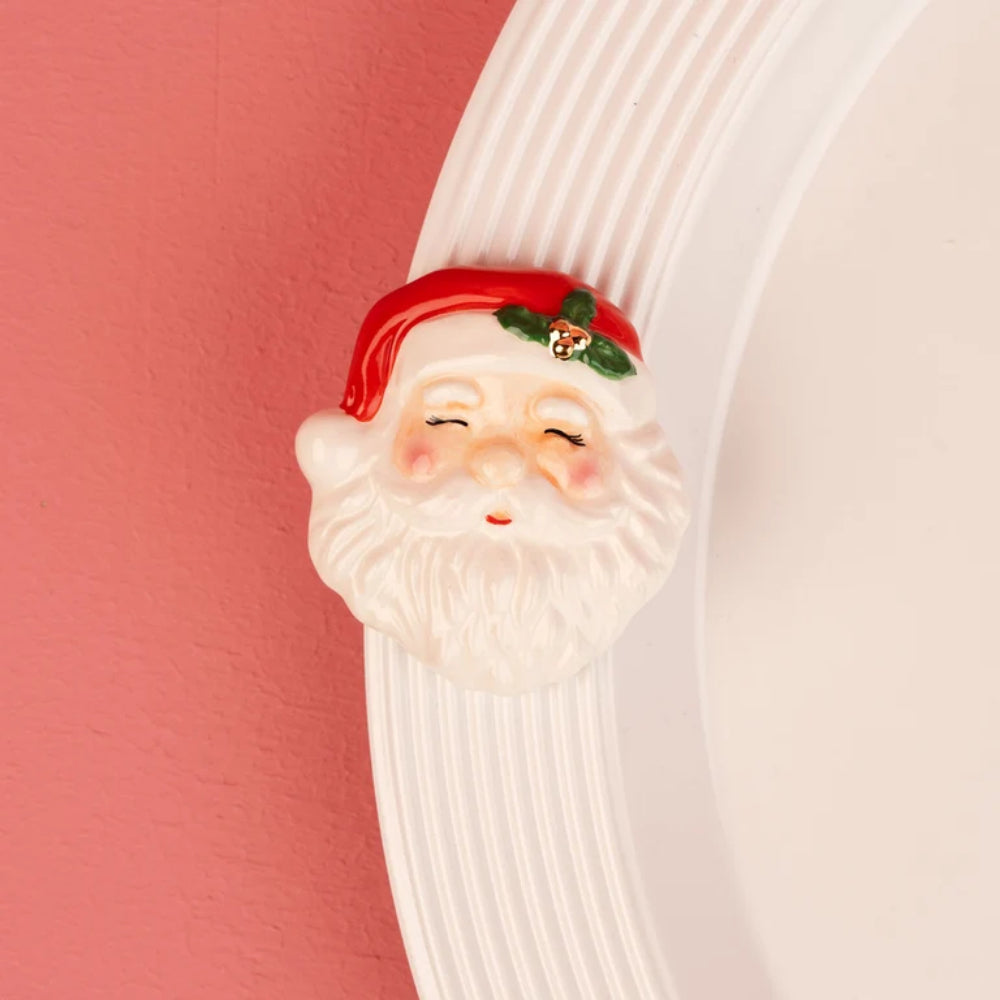 A ceramic Santa Claus head wall hook with a red hat, green holly accent, and white beard, shown mounted on a white ribbed wall next to a pink wall.