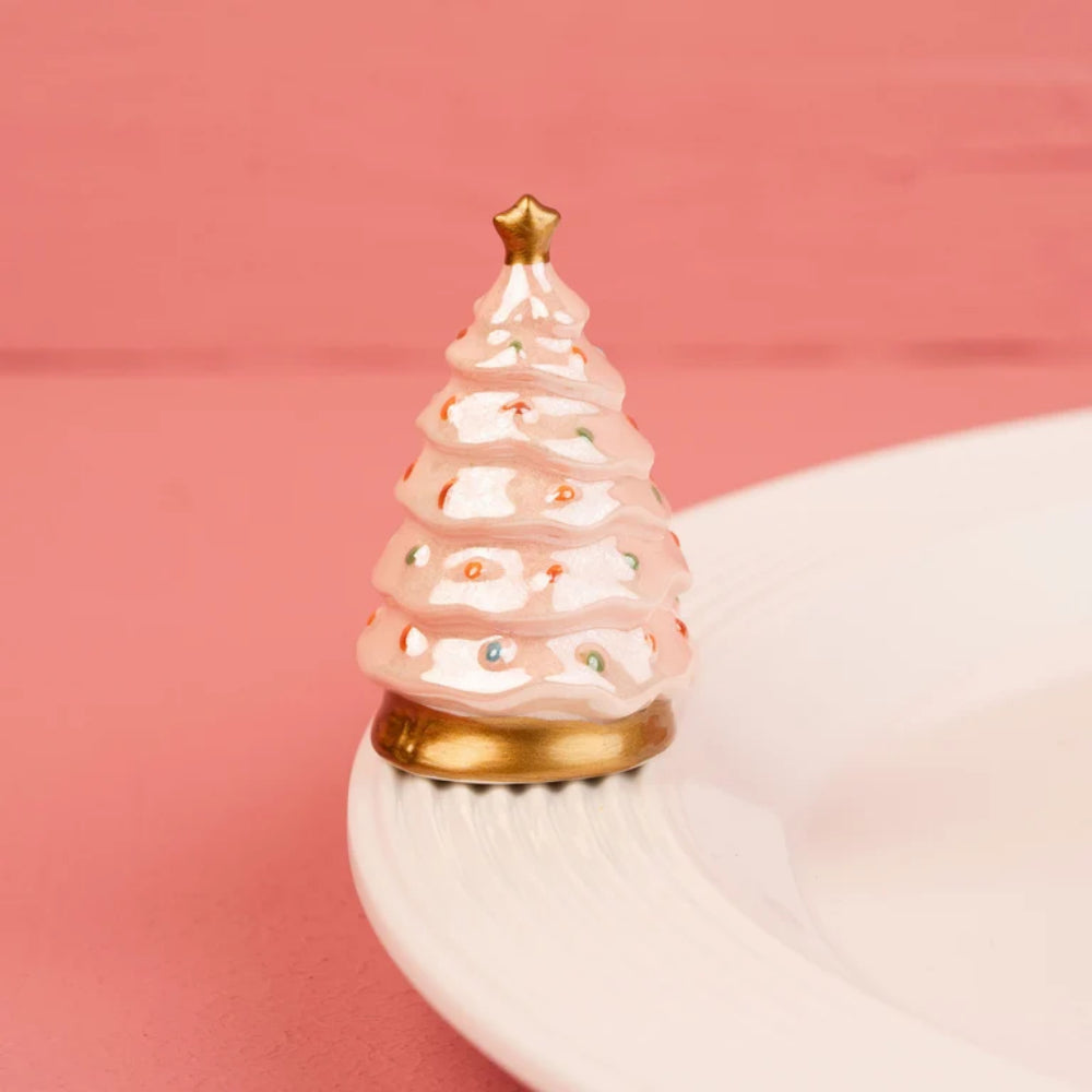 A small decorative Christmas tree figurine with a pink and white frosting-like textured body, gold star topper, and gold base, displayed on a white plate against a pink background.