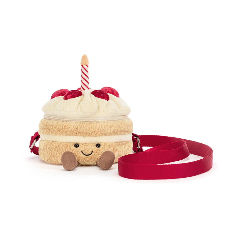 A plush cake-shaped crossbody bag with a cream-colored body, red frosting, a red-and-white striped candle, and a red strap, photographed on a white background.
