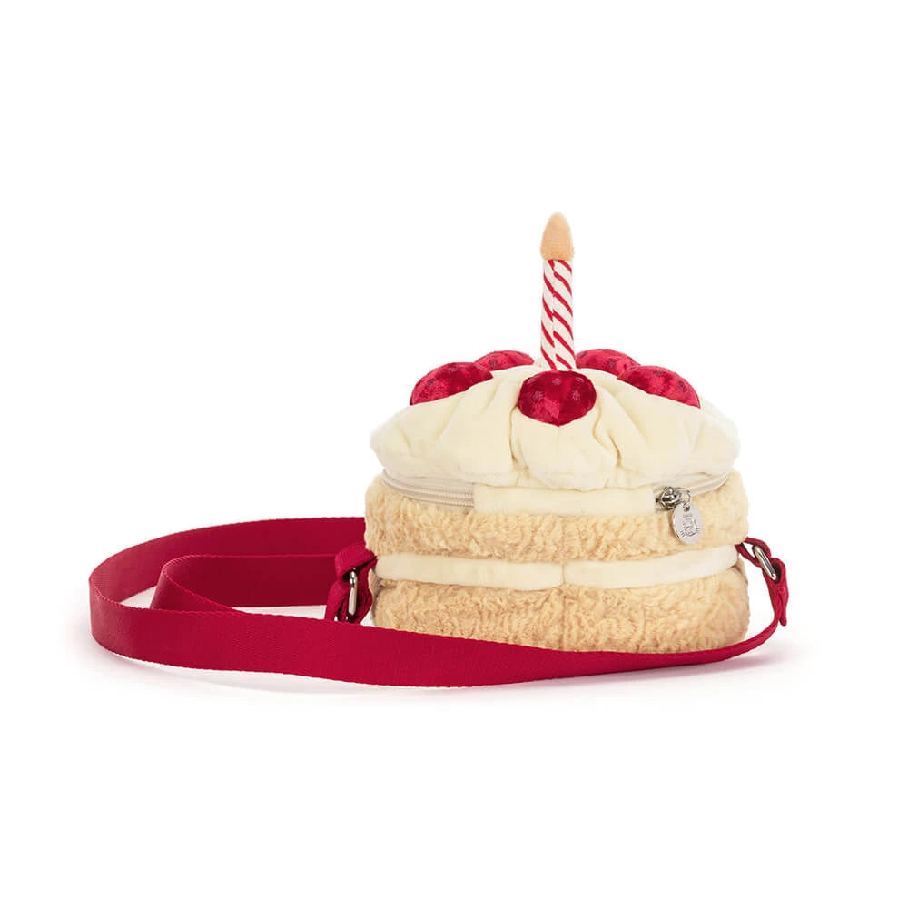 A plush cake-shaped crossbody bag with cream-colored faux frosting, red cherry details, a red ribbon, and a single red-and-white striped candle on top, shown on a white background.