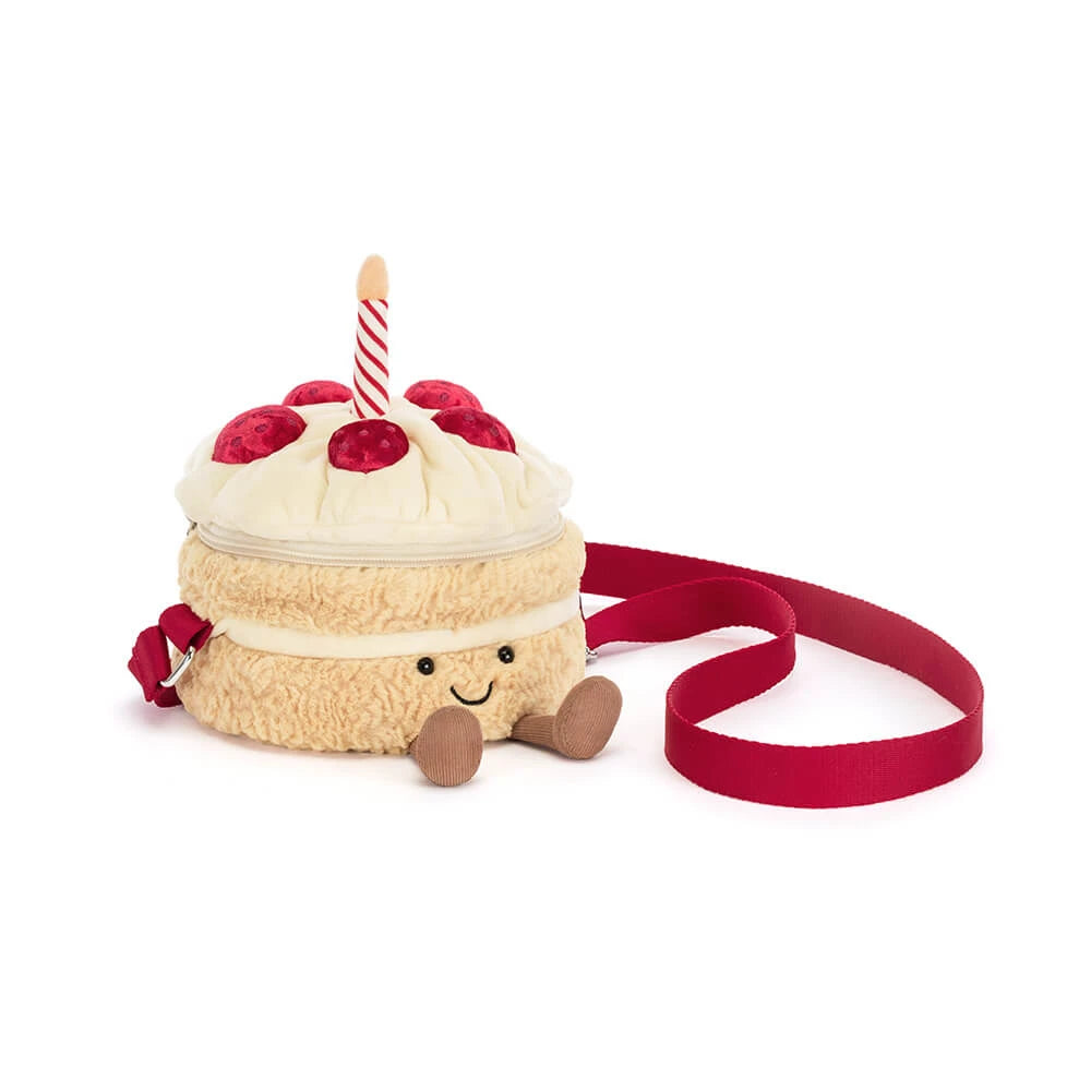 A plush cake-shaped crossbody bag with a red ribbon strap, featuring cream frosting, red strawberries, a candle, and a smiling face on a white background.