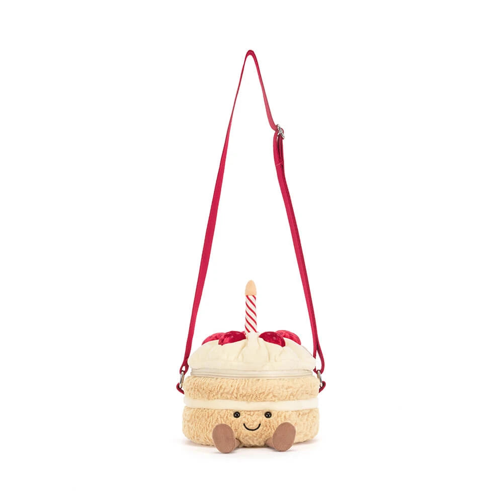 A plush cake-shaped shoulder bag with a cream-colored fuzzy body, red frosting details, a red and white striped candle, and a red adjustable strap, shown on a plain white background.