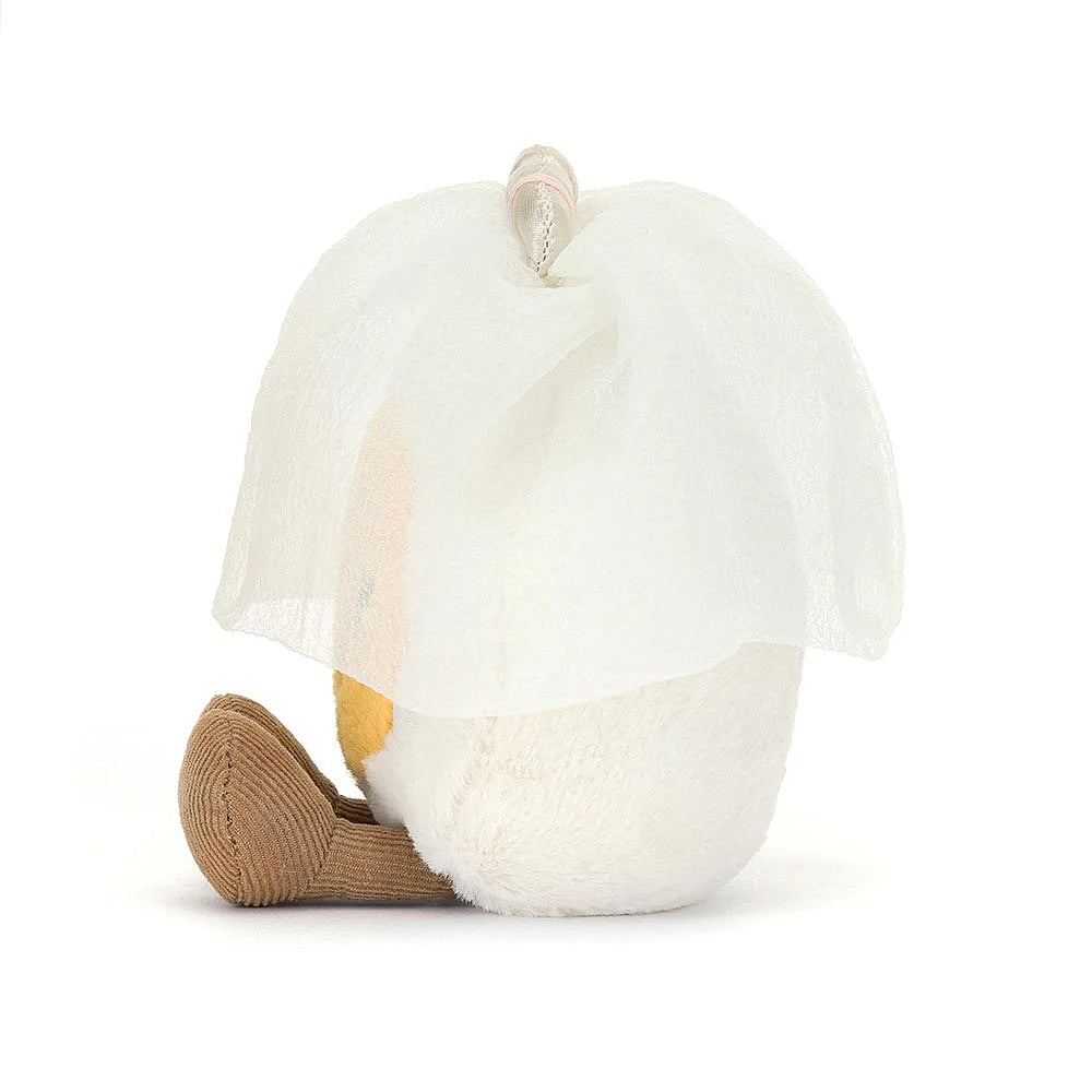 A side photo of a plush toy representing a boiled egg dressed as a bride, with a white veil and a yellow face, sitting upright with cocoa cordy legs.