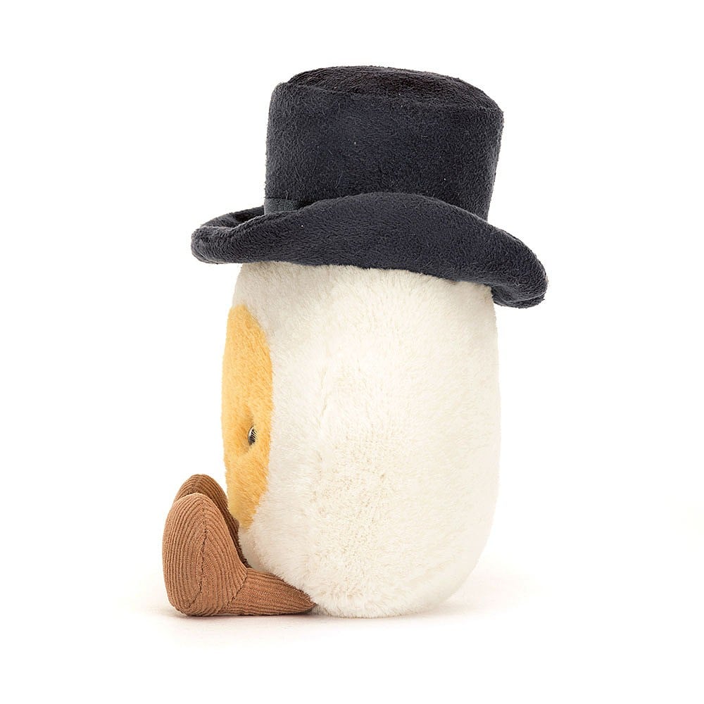 A side photo the  of a plush toy representing a boiled egg dressed as a groom with a black hat and  yellow face, sitting upright with cocoa cordy legs.