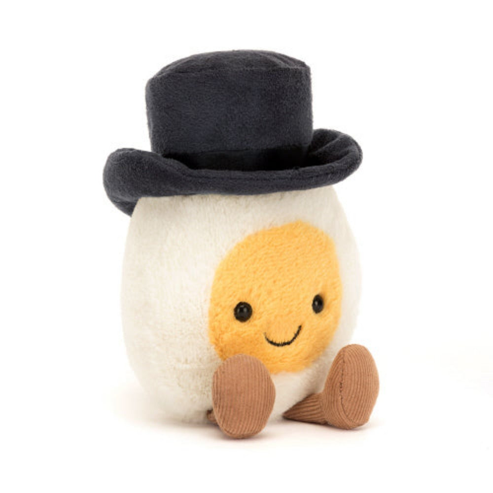 A angled photo of a plush toy representing a boiled egg dressed as a groom with a black hat and  yellow face, sitting upright with cocoa cordy legs.