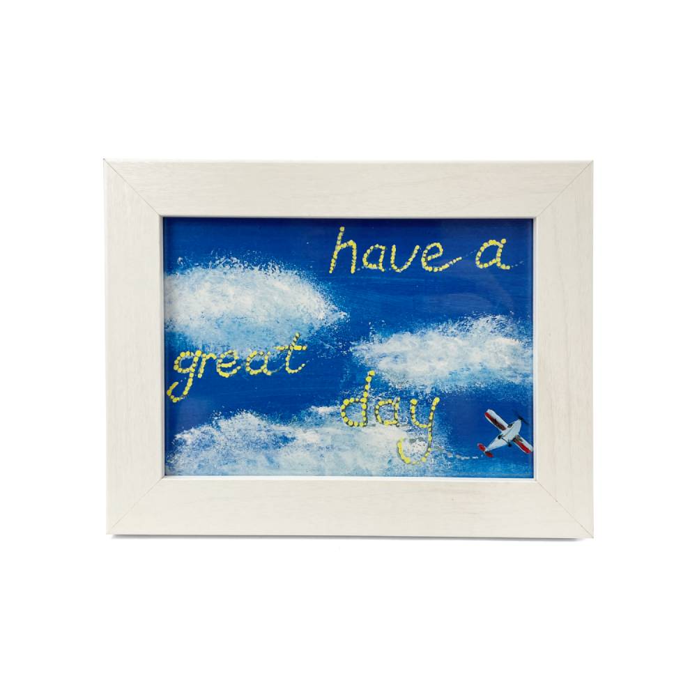 A framed art print featuring a blue sky with white clouds, yellow glittery text reading 'have a great day', and a small red-and-white airplane, set in a light wood frame against a white background.
