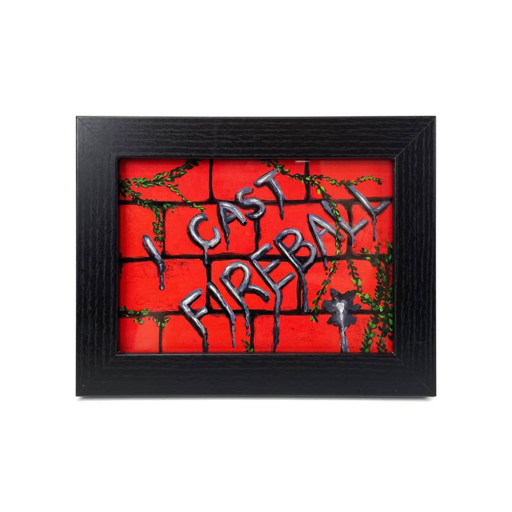 A framed art piece featuring a red brick wall background with the words 'I CAST FIREBALL' in dripping metallic letters, accented with green ivy, enclosed in a black textured frame.