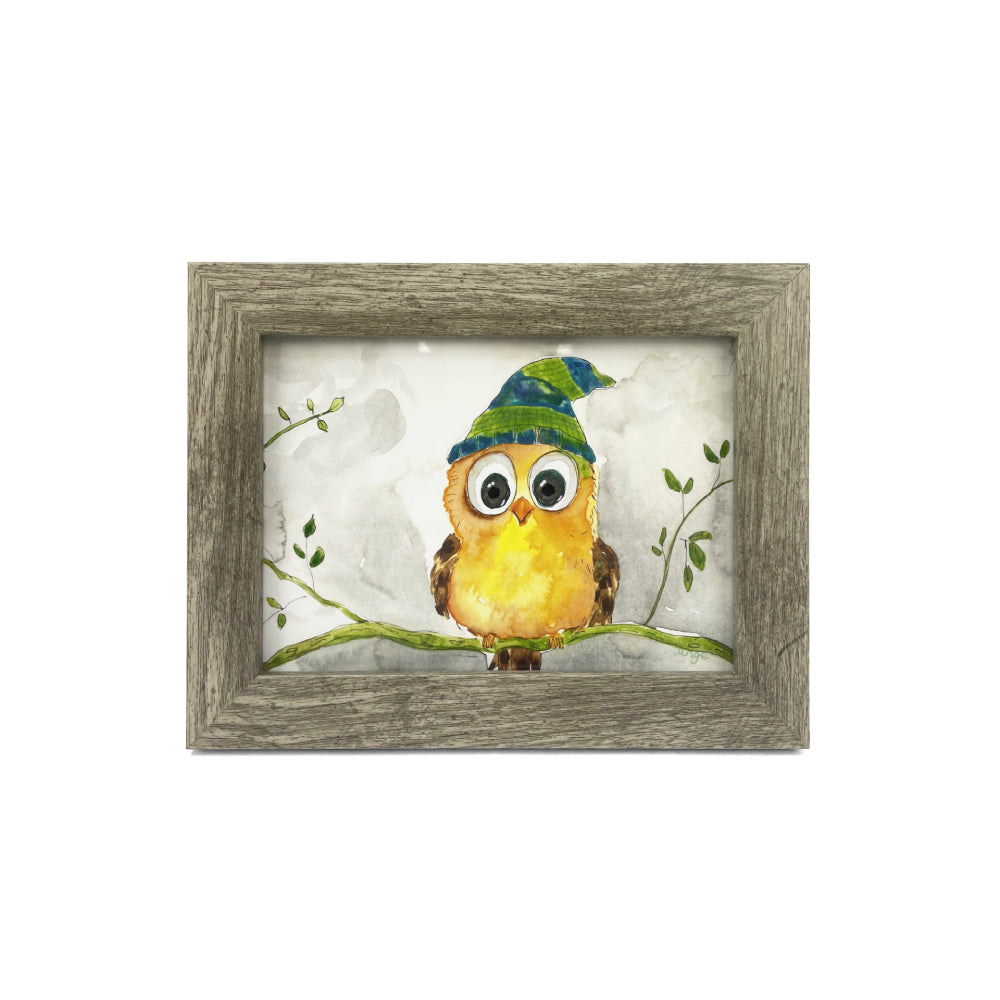 Framed artwork of an yellow owlet wearing a green beanie on a branch with leaves.