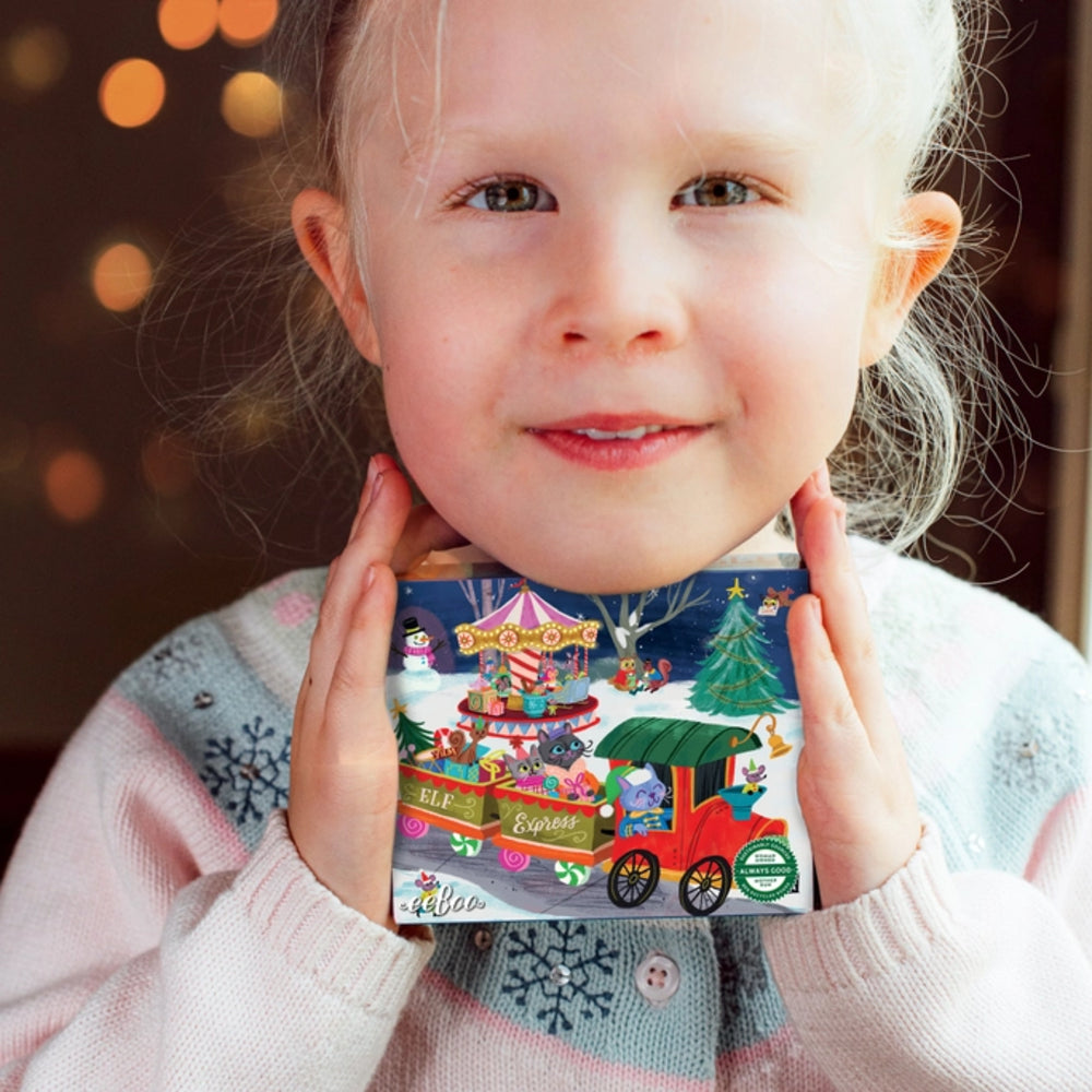A young child in a light pink knit sweater holds a colorful illustrated advent calendar box in front of their face. The box features a winter scene with a red train, Christmas tree, market stall, and snow, with the text 'A Christmas Countdown' and 'Mila' visible.