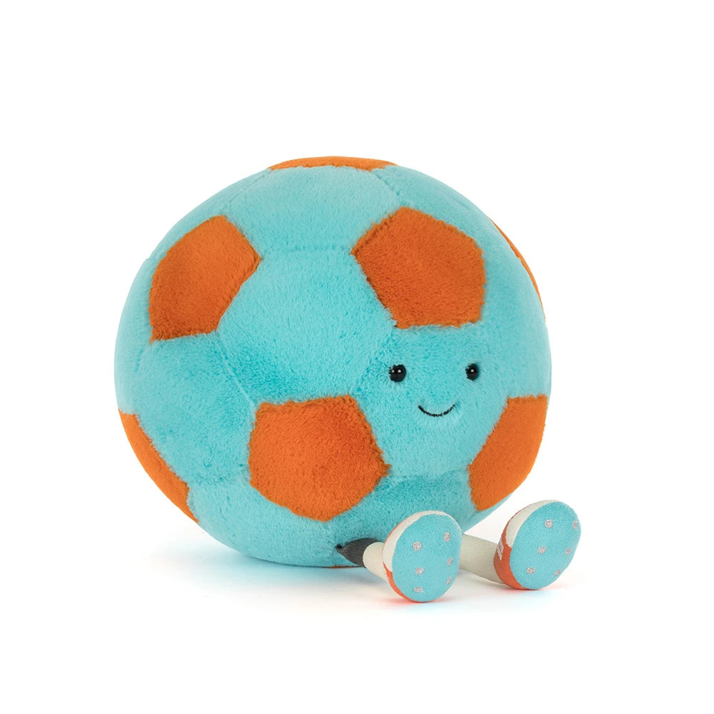 A side angled photo of a teal and orange Jellycat Amuseables Soccer Ball push toy with a smiling face and white legs on a white background.