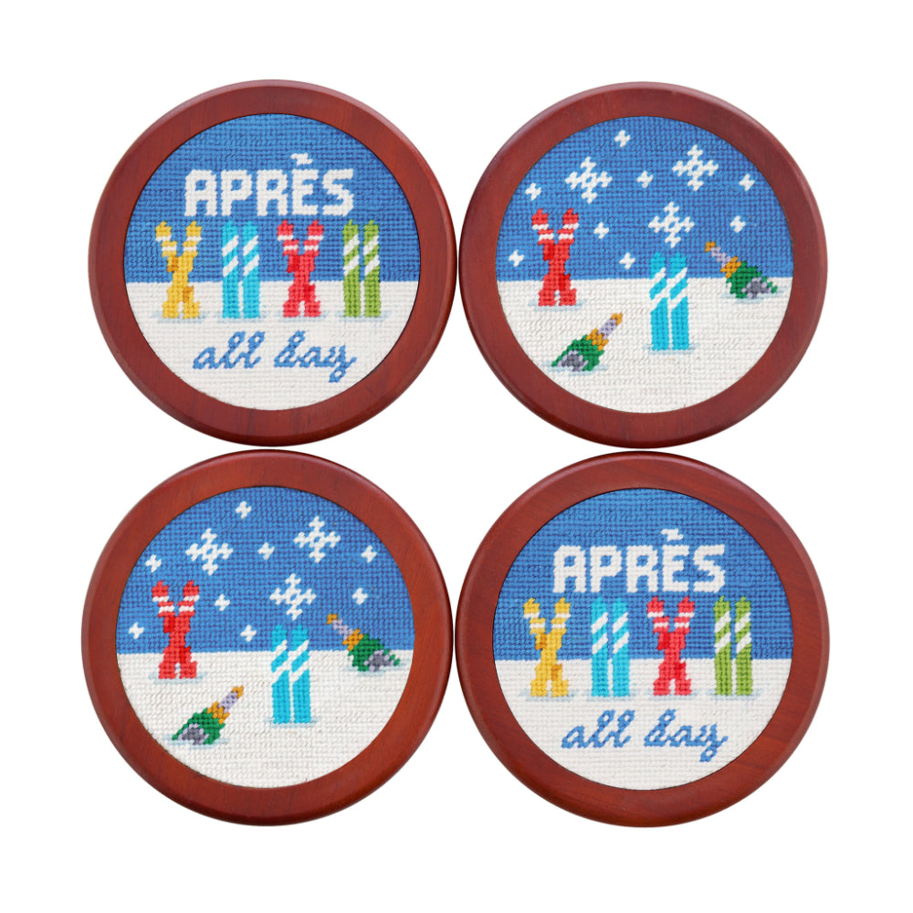 Four round wooden coasters arranged in a 2x2 grid on a white background. Each coaster features a cross-stitch style winter scene with snow, colorful skis, and the words “APRÈS all day” in blue.