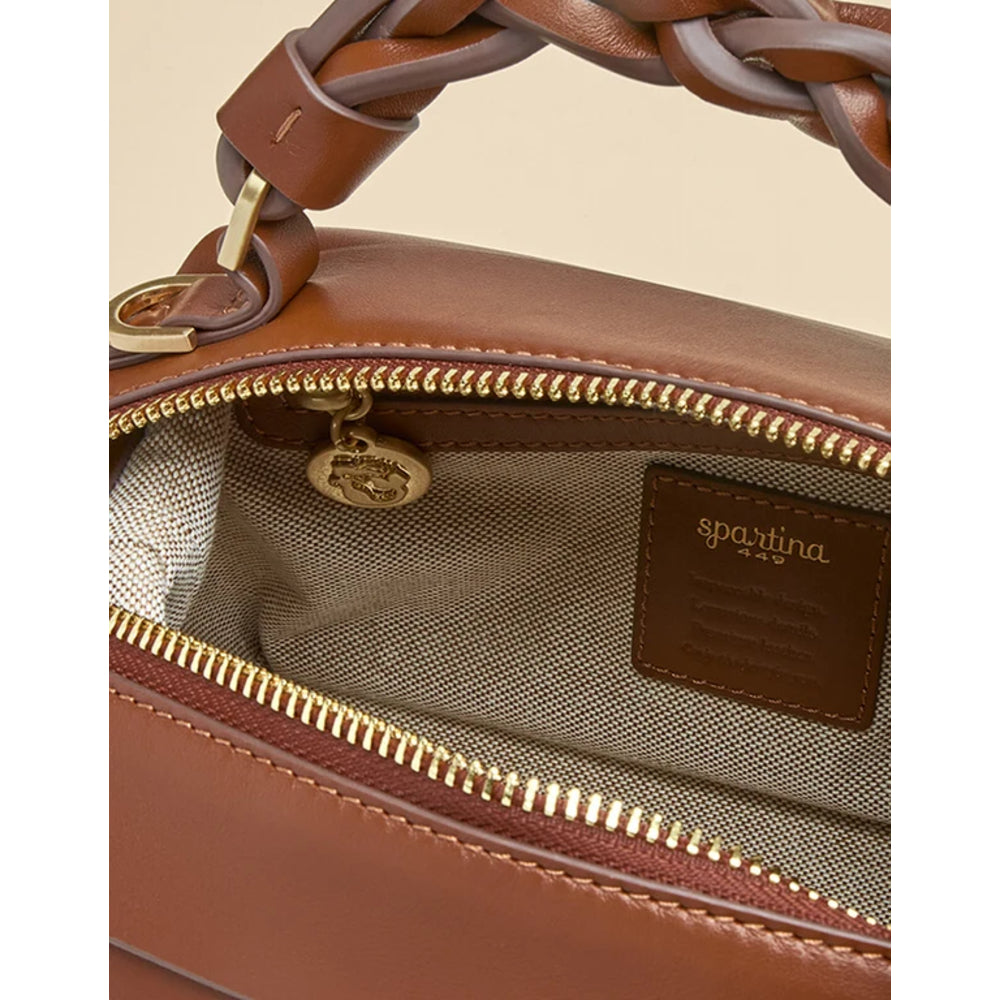 Close-up of a brown leather handbag interior with a gold zipper, a brown leather brand patch reading 'spartina', and part of a braided top handle visible against a light beige background.