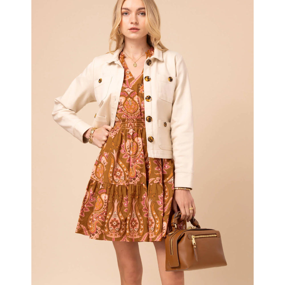 A woman models a cream-colored denim jacket over a brown and pink paisley tiered dress, holding a brown leather handbag against a solid beige background.