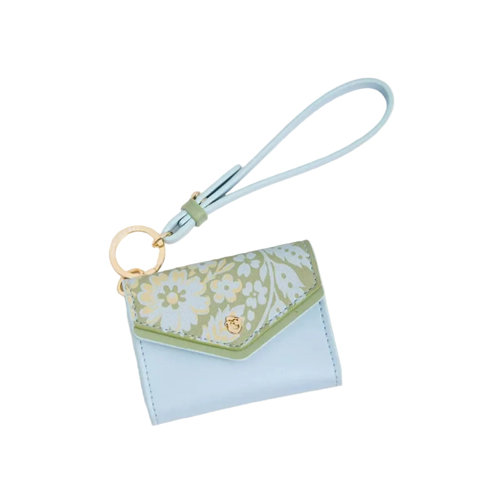 A light blue leather wristlet wallet with a floral-patterned flap in sage green, cream, and light blue, featuring a gold-tone ring and a matching light blue wrist strap, set against a solid white background.