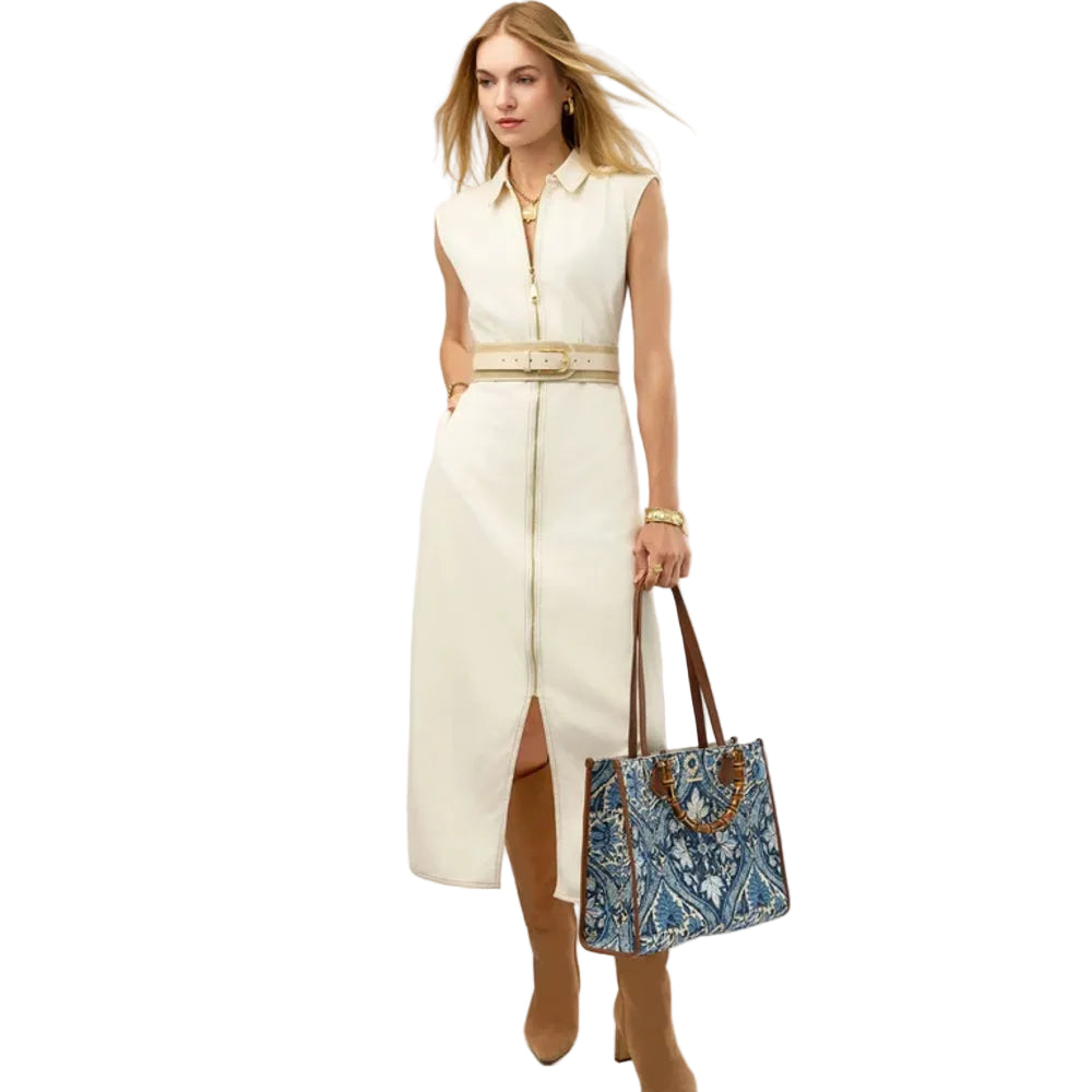 A woman models a sleeveless cream midi dress with a front zipper, collar, and matching belt. She is holding a blue and white patterned tote bag with brown handles, set against a solid white studio background.