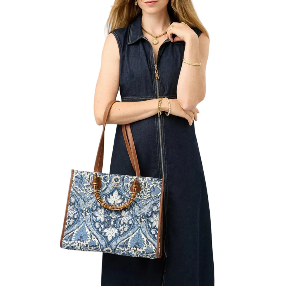 A woman wearing a dark navy sleeveless zip-front dress holds a blue and white patterned tote bag with brown leather handles and trim against a plain light beige background.