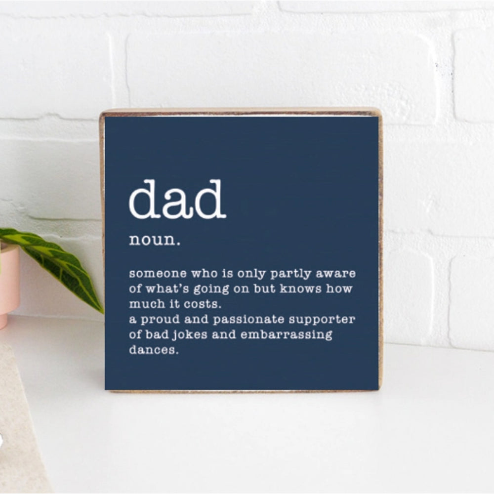 Navy blue wood  square block sign with white text definition of 'dad'  on a white kitchen counter with a white brick wall 
