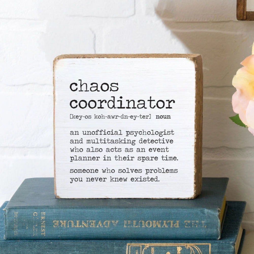 White wood decorative square block sign with the definition of 'chaos coordinator'  "An Unofficial Psychologist And Multitasking Detective Who Also Acts As An Event Planner In Their Spare Time. Someone Who Solves Problems You Never Knew Existed" in black text on top of books with a white wall in the backround 