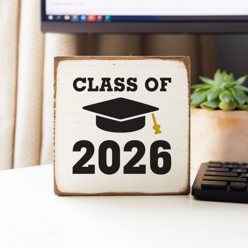 White wood cubed shaped decorative   square block sign with 'Class of 2026' in black text in on a desk with a computer and plant in the background