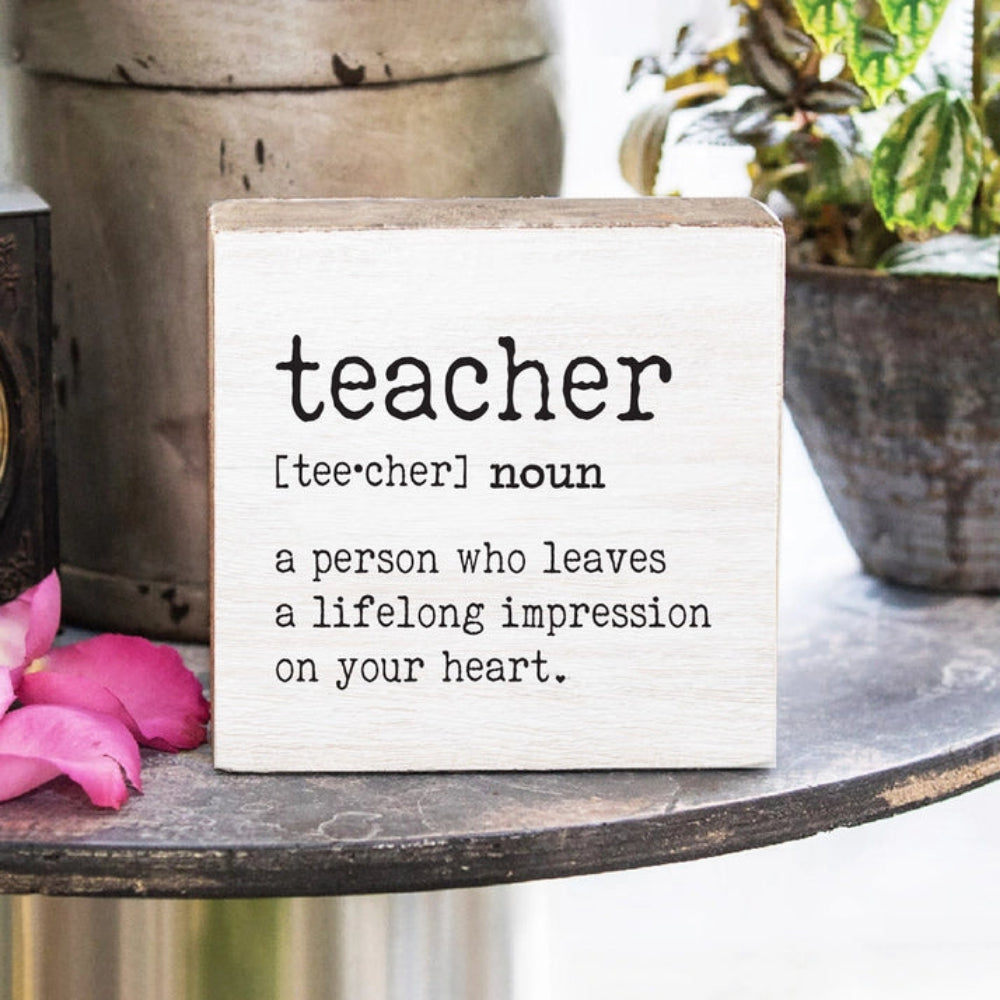  White wood decorative square block sign with definition of 'teacher' "a person who leaves a lifelong impression on your heart" in black text on a table with flowers and plants.