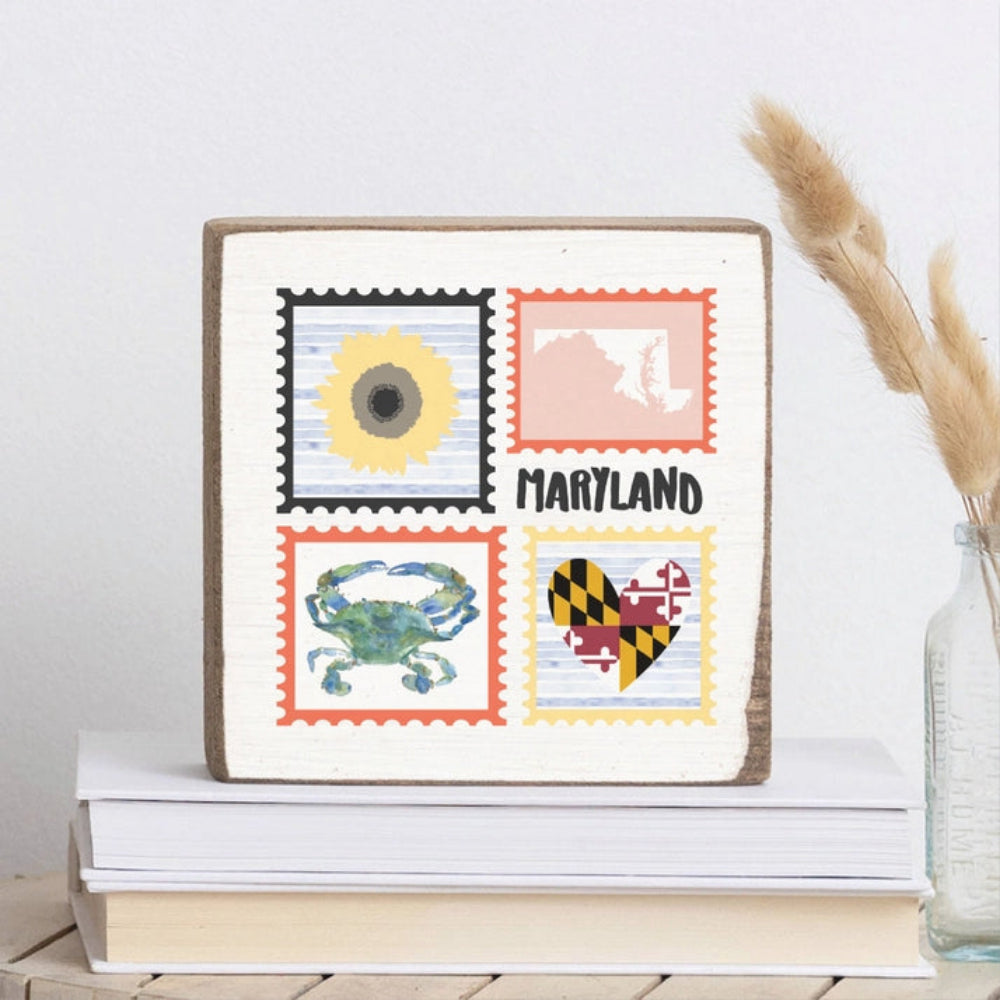A white wood  decorative square  block sign with postage stamp designs featuring a sun, state outline, crab, and Maryland flag, with 'Maryland' text on two books with a clear vase with a beige plant decor in it 
