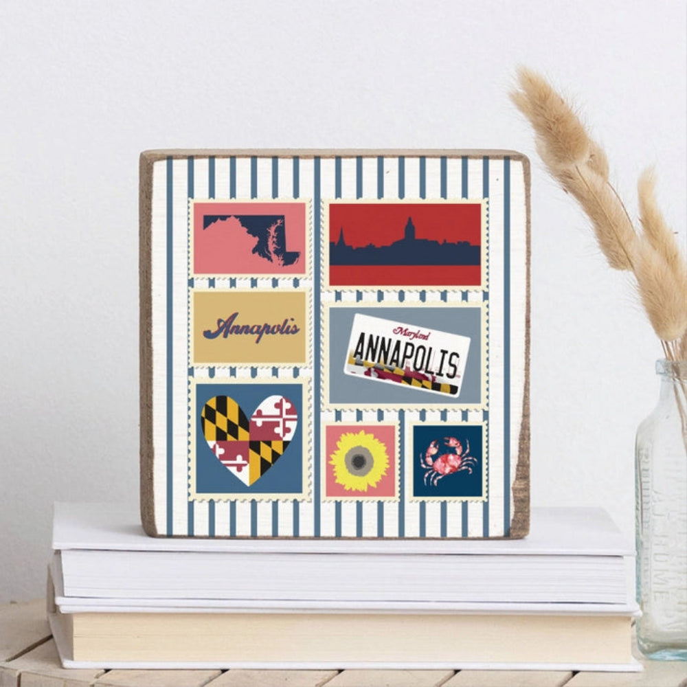 White and blue striped wood decorative square block sign with maryland-themed postage stamps featuring icons and text about annapolis, maryland , placed on books with a clear vase with beige  dried plants on a grey backround 