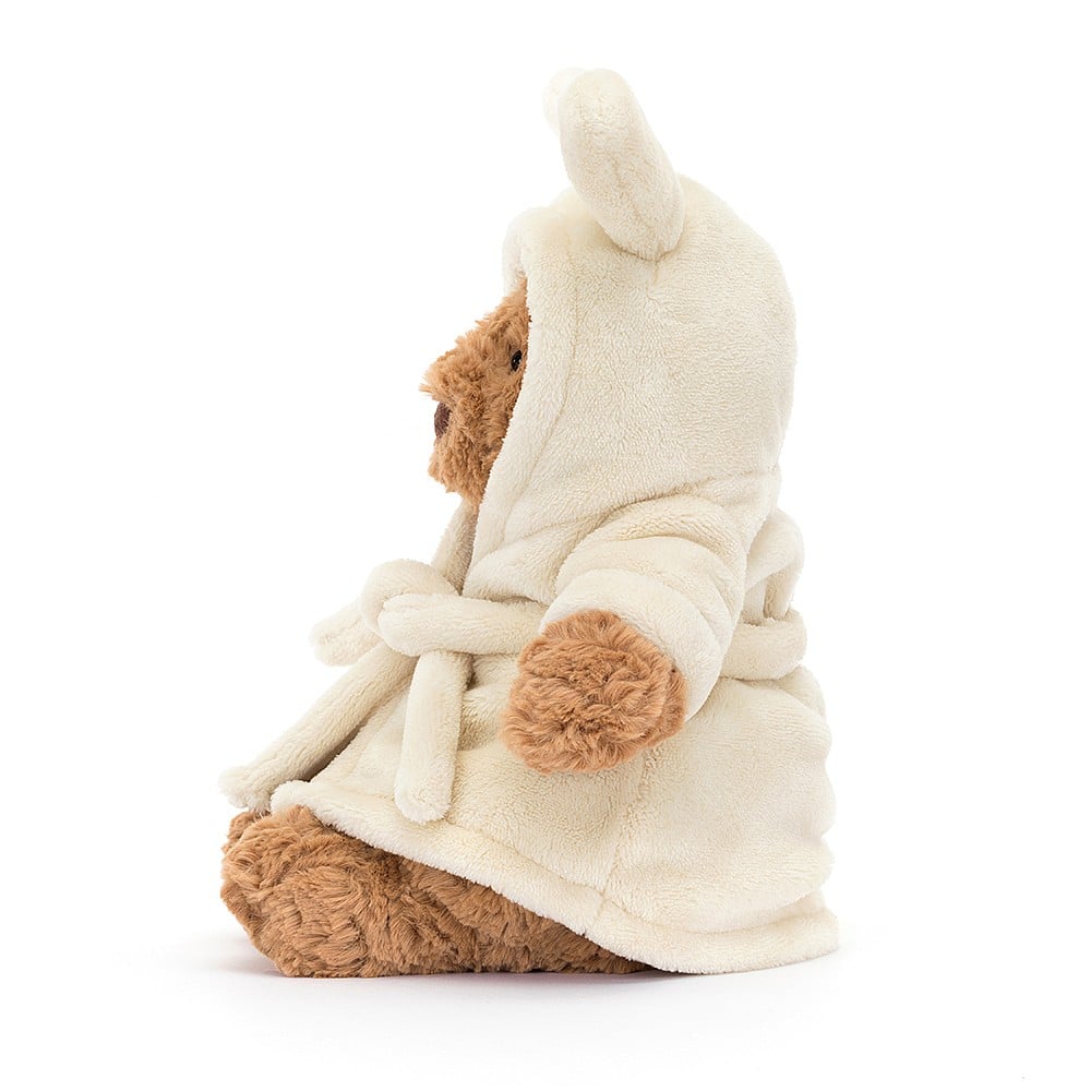A side photo of an stuffed teddy bear wearing a cream bath robe with a hood and bunny ears on a white background.