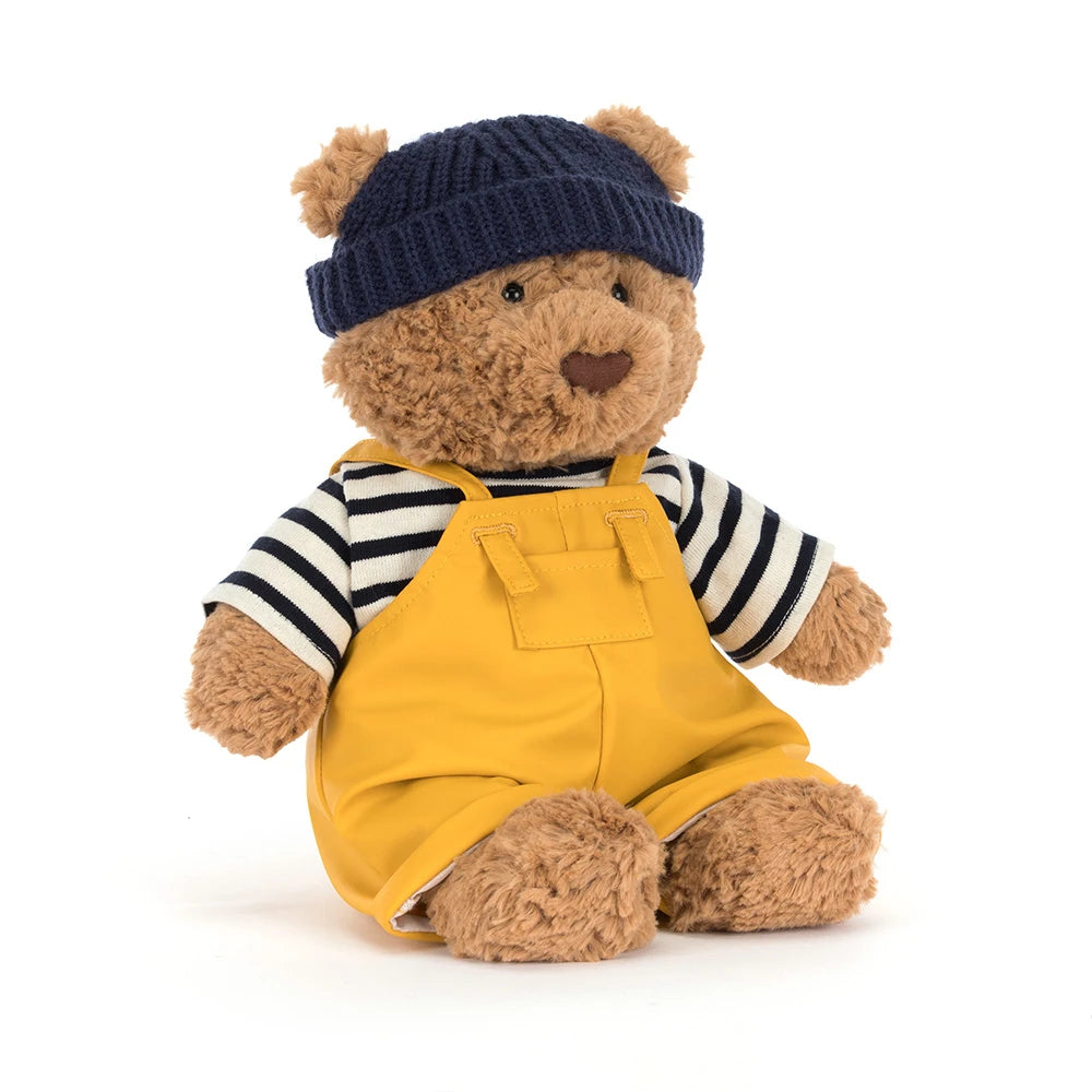 Angled side photo of a beige teddy bear with a colorful seafarer outfit with yellow overalls,navy cap and a white and blue striped shirt on a white backround