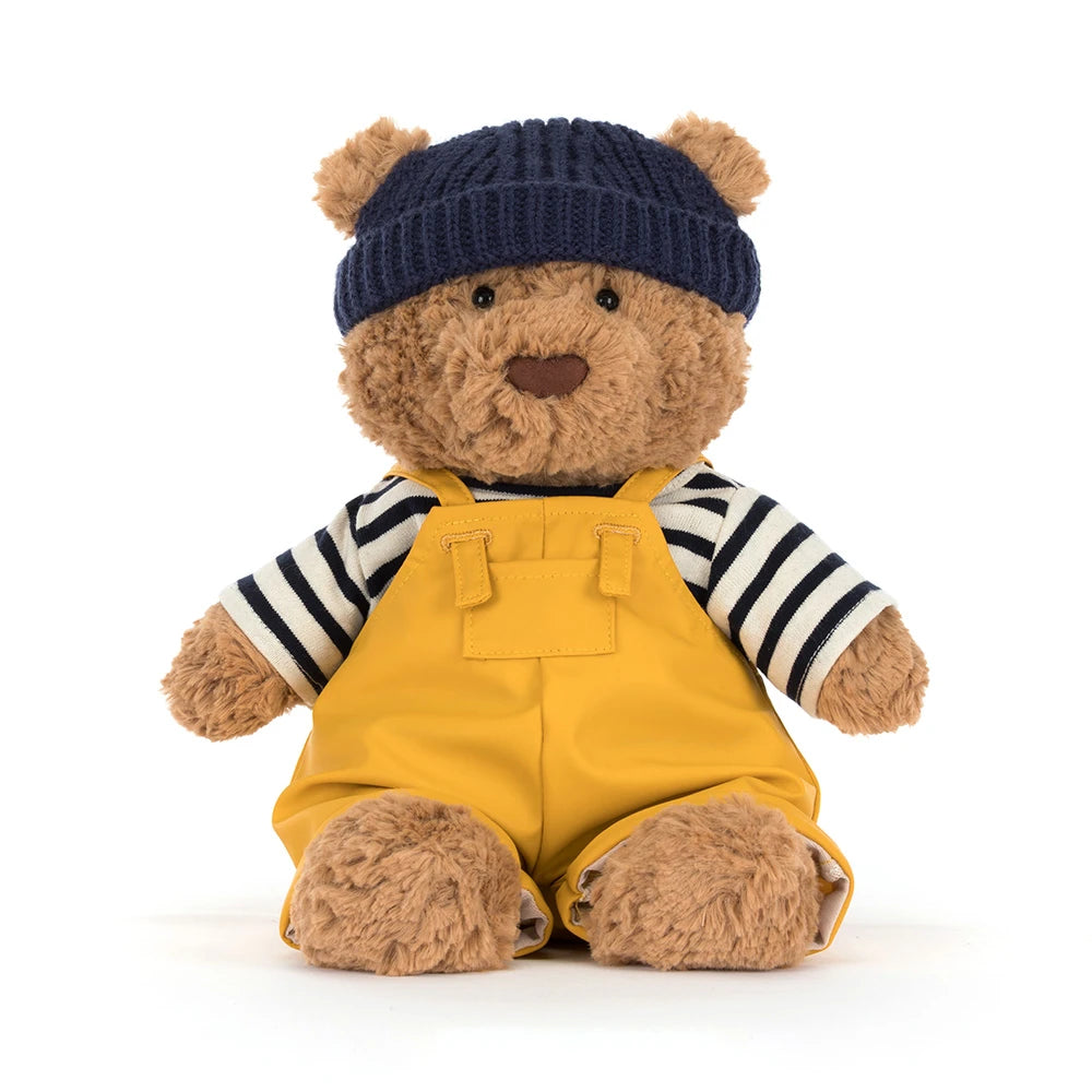 Beige teddy bear with a colorful seafarer outfit with yellow overalls,navy cap and a white and blue striped shirt on a white backround