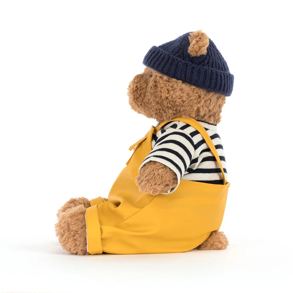 A side photo of a beige teddy bear with a colorful seafarer outfit with yellow overalls,navy cap and a white and blue striped shirt on a white backround