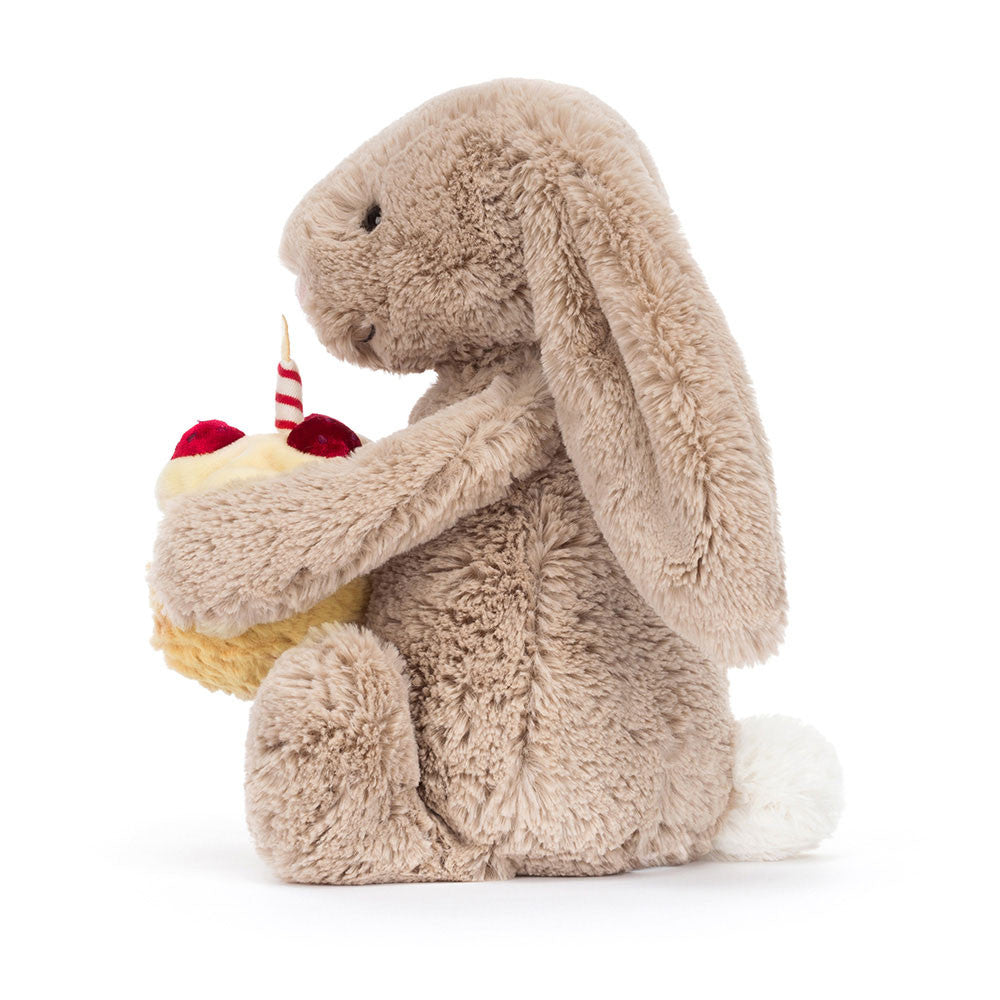 A side photo of a  beige plush toy bunny holding a cupcake with a candle on a white background.