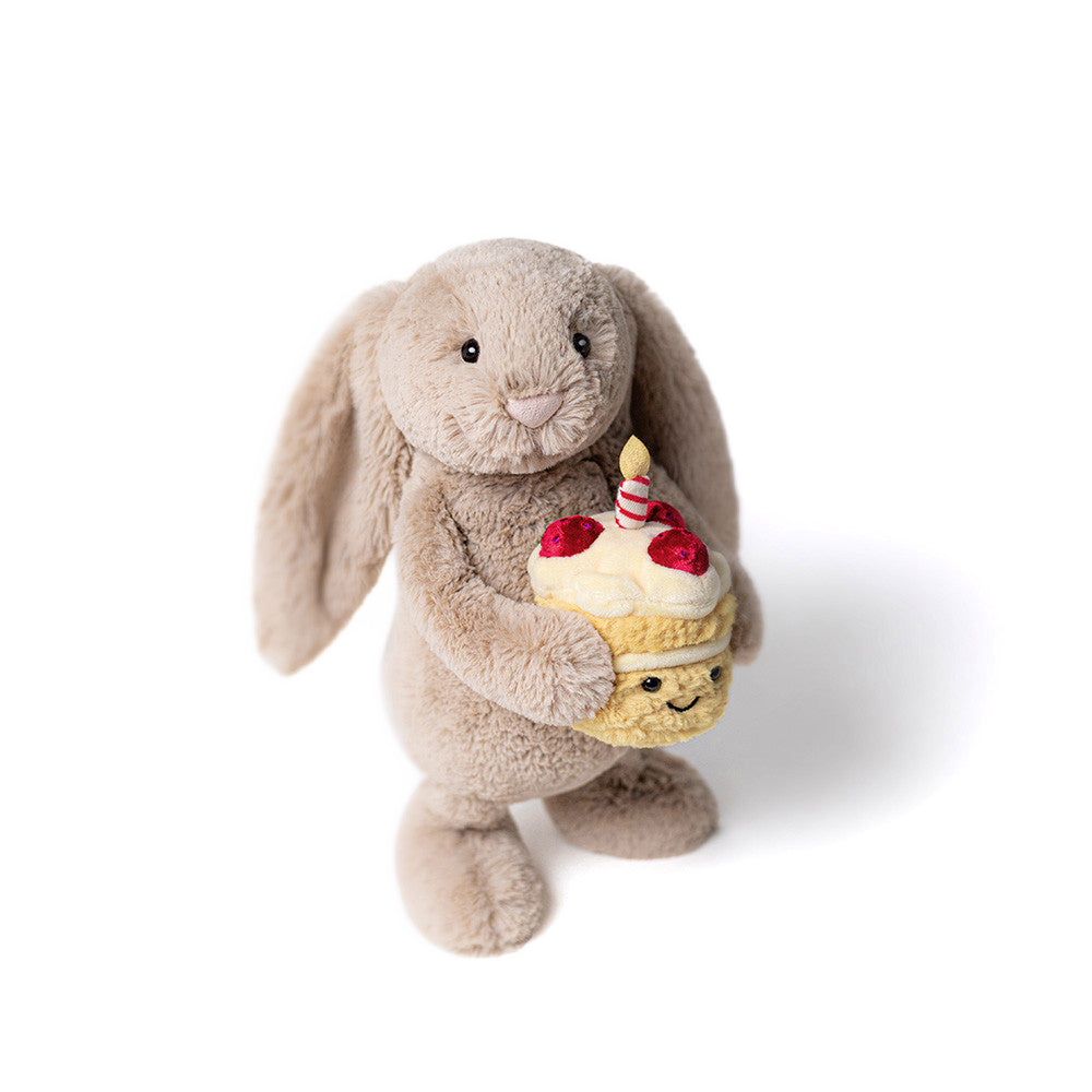 A beige plush toy bunny standing holding a cupcake with a candle on a white background