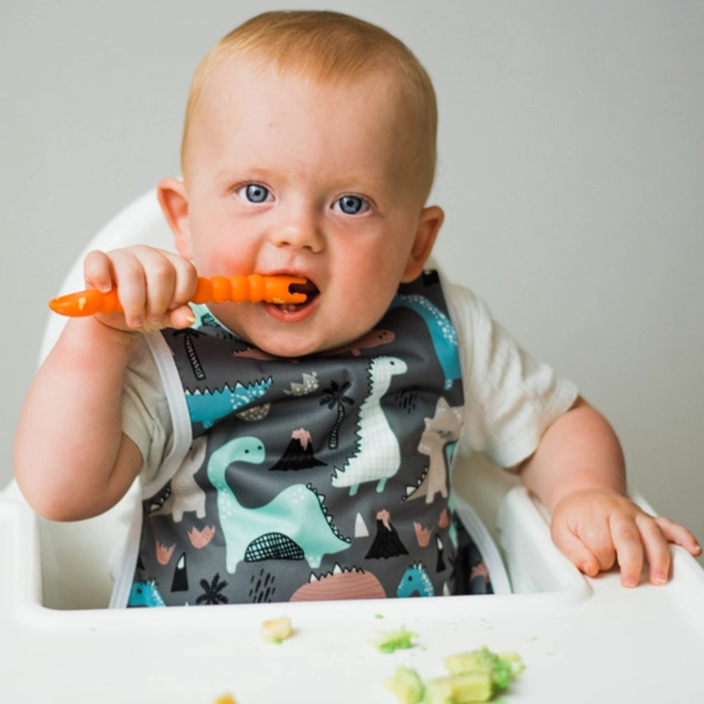 Baby wearing a dinosaur-patterned bib while eating with an orange spoon.