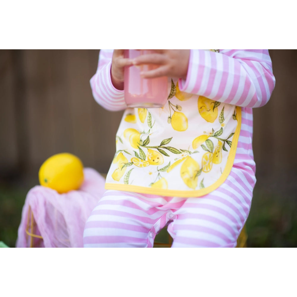 Child wearing a pink and white striped outfit with a lemon-patterned bib, holding a pink cup outdoors.