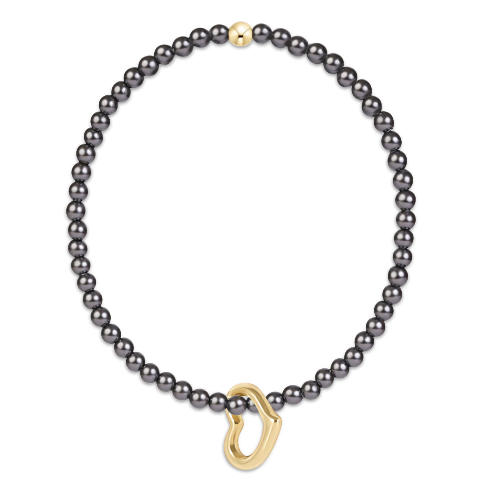 A delicate bracelet featuring  small, dark gray pearls and a polished gold-tone open heart charm, displayed on a clean white background.
