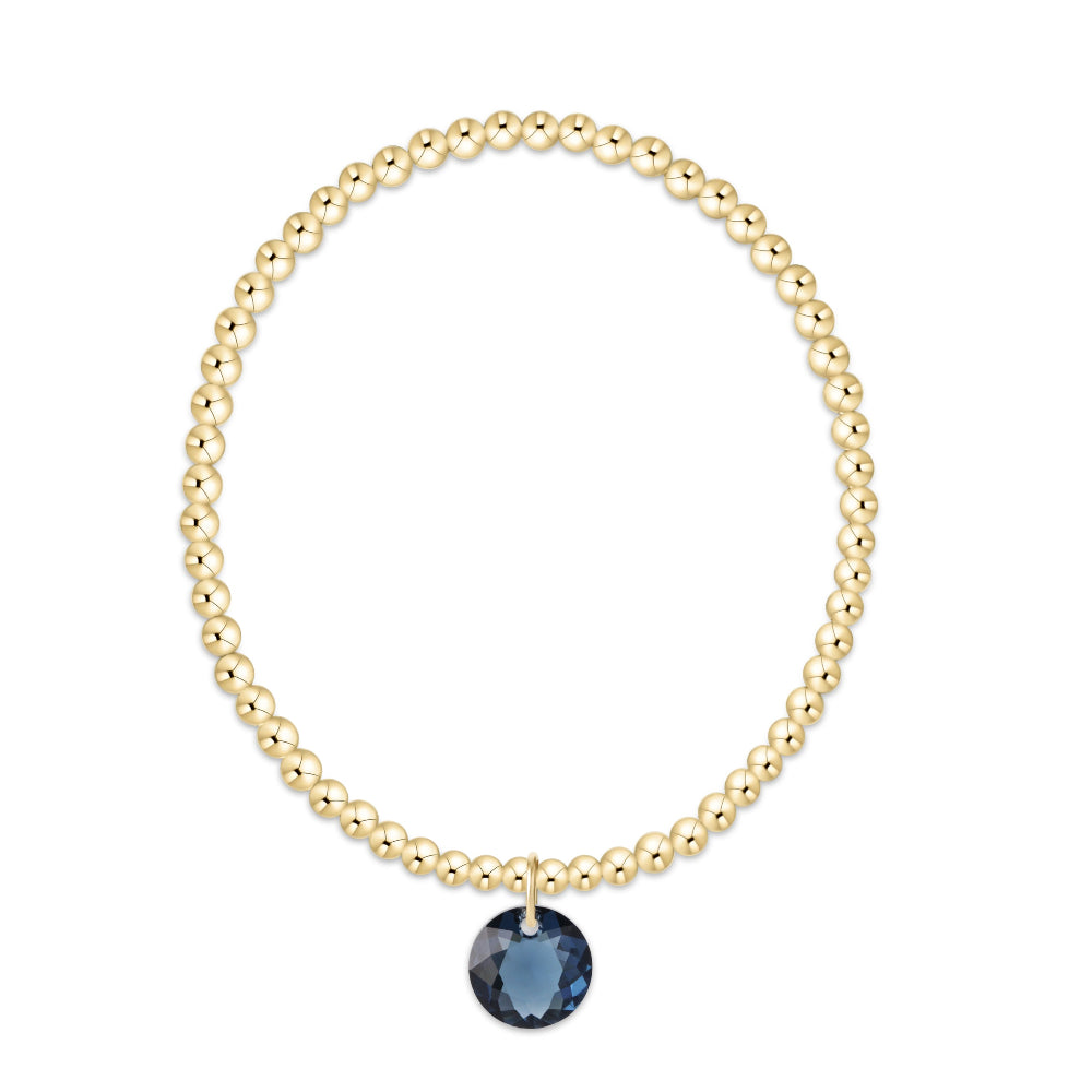 A gold beaded bracelet with a single faceted dark blue gemstone pendant, displayed on a clean white background.