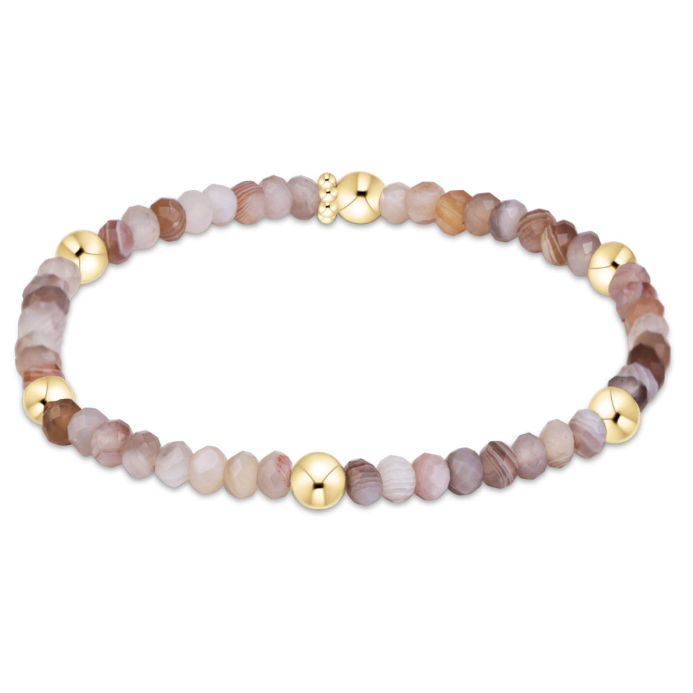 A beaded stretch bracelet featuring faceted natural stone beads in shades of brown, beige, and lavender, accented by polished gold-tone metal beads and a clasp, shown on a plain white background.
