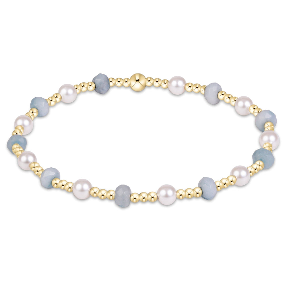A delicate beaded bracelet featuring light blue-gray beads, white pearls, and polished gold-tone spacer beads, arranged in an oval shape on a clean white background.