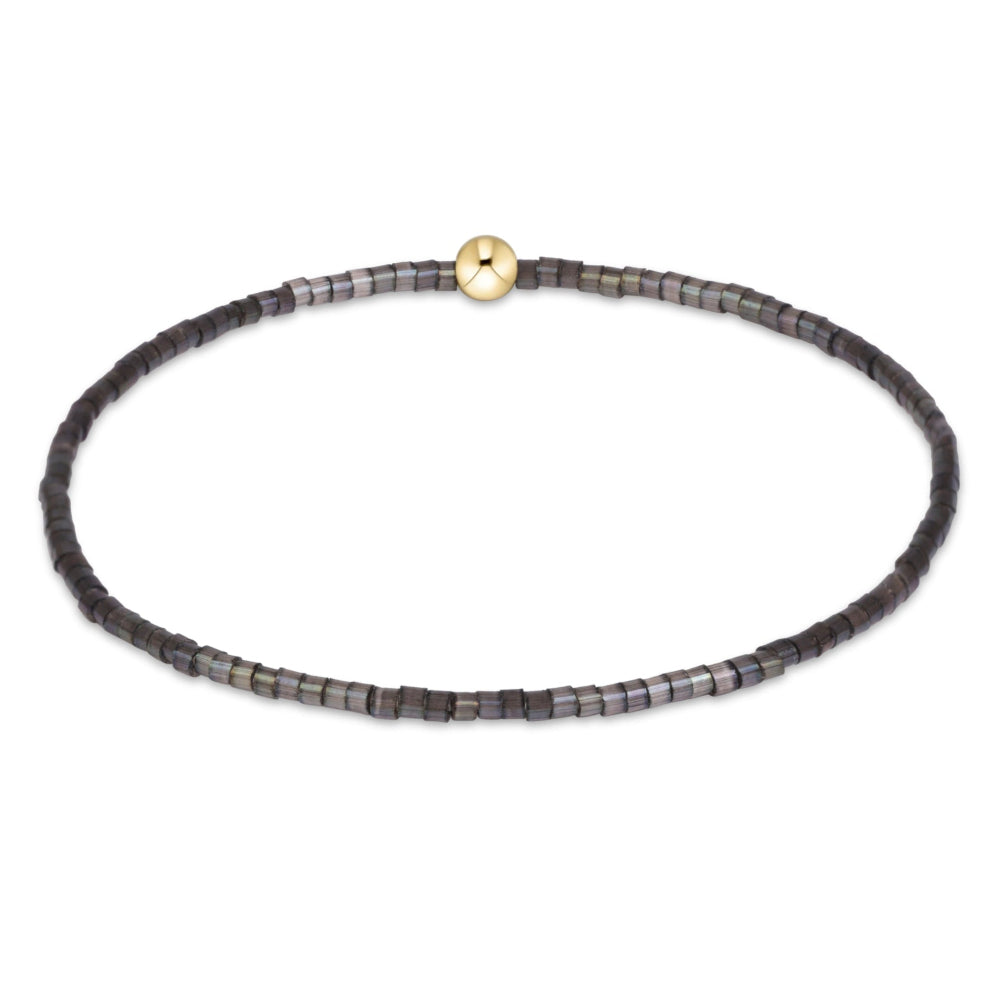 A beaded stretch bracelet featuring small dark gray and black square beads with a shimmering iridescent finish, accented by a single round gold-tone bead at the top center, shown on a plain white background.
