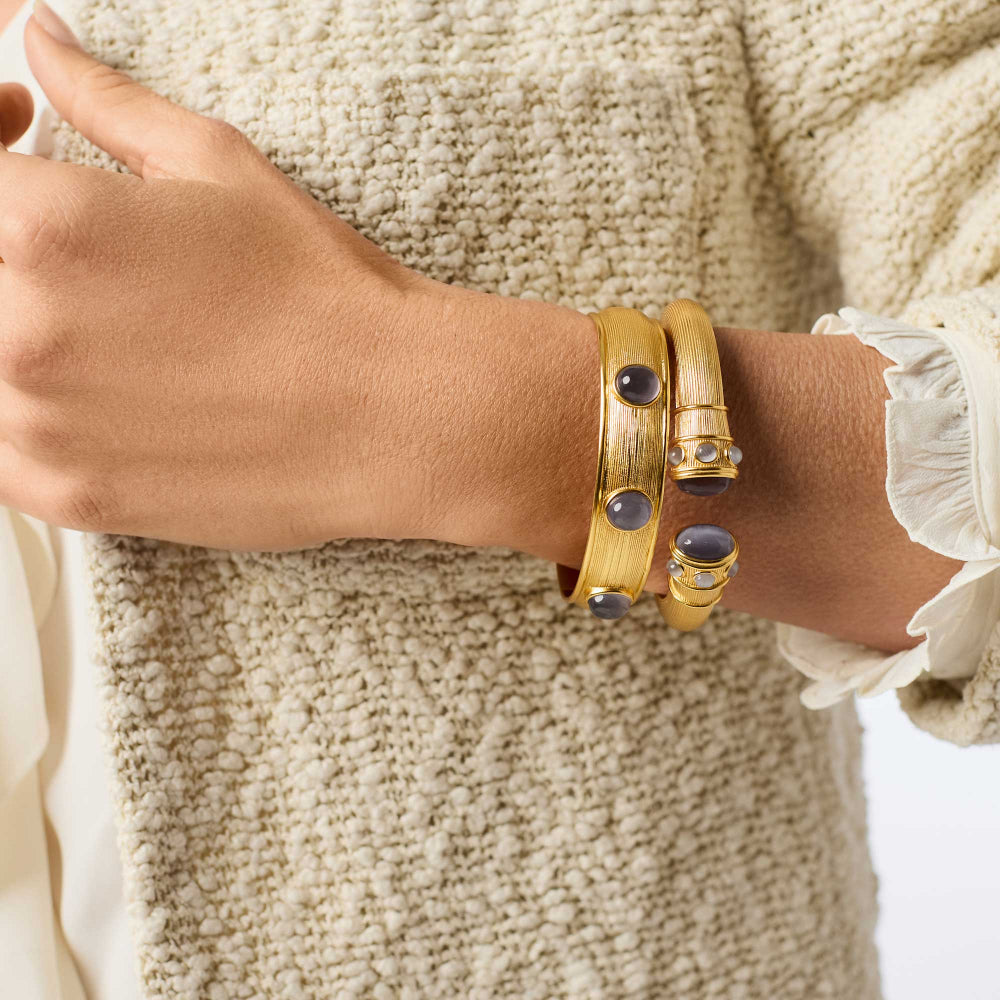 A close-up of a person's wrist wearing three stacked gold-toned bracelets, including one with dark oval stones and two with gold-toned cylindrical links and clear stone accents, against a cream textured sweater.
