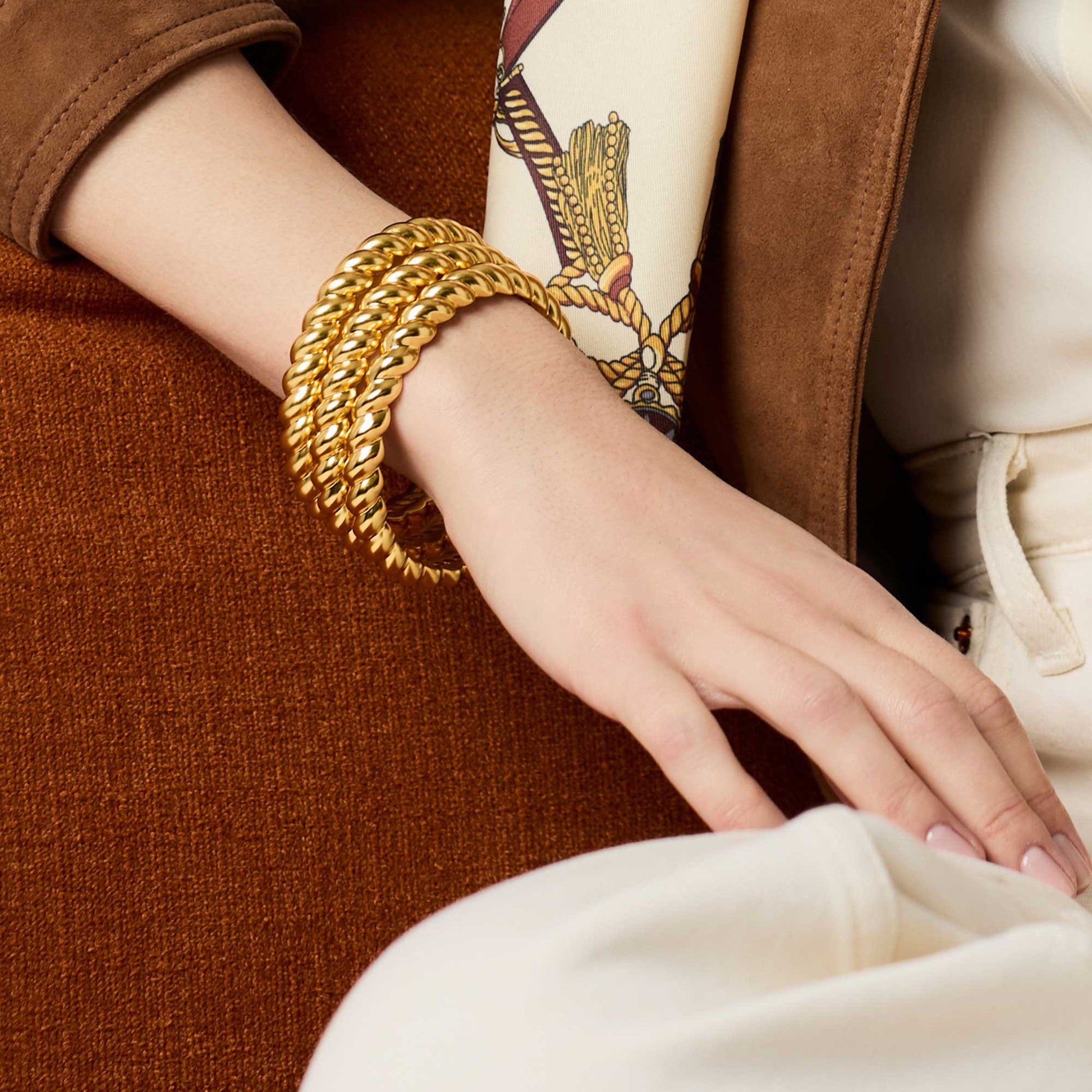 Close-up of a hand wearing gold bracelets with a brown coat and patterned scarf in the background