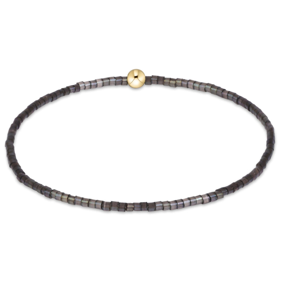 A beaded stretch bracelet featuring small dark gray and black square beads with a shimmering iridescent finish, accented by a single round gold-tone bead at the top center, shown on a plain white background.