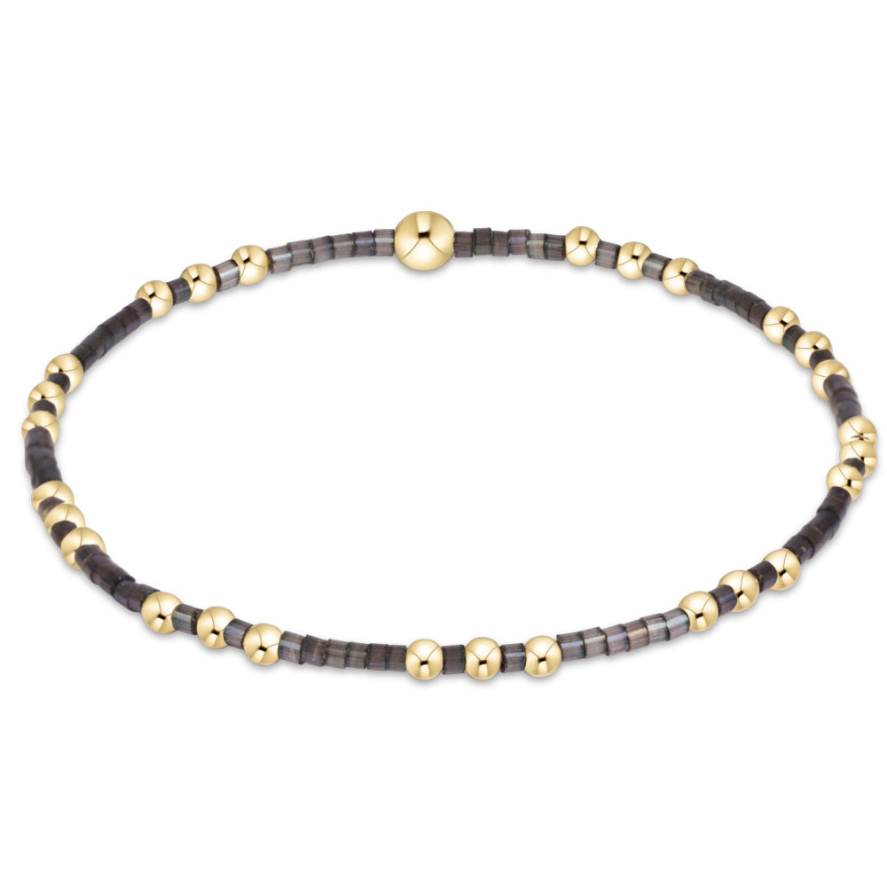 A delicate beaded stretch bracelet featuring alternating dark gray/black beads and small gold spacer beads, with a polished gold bead, shown on a plain white background.