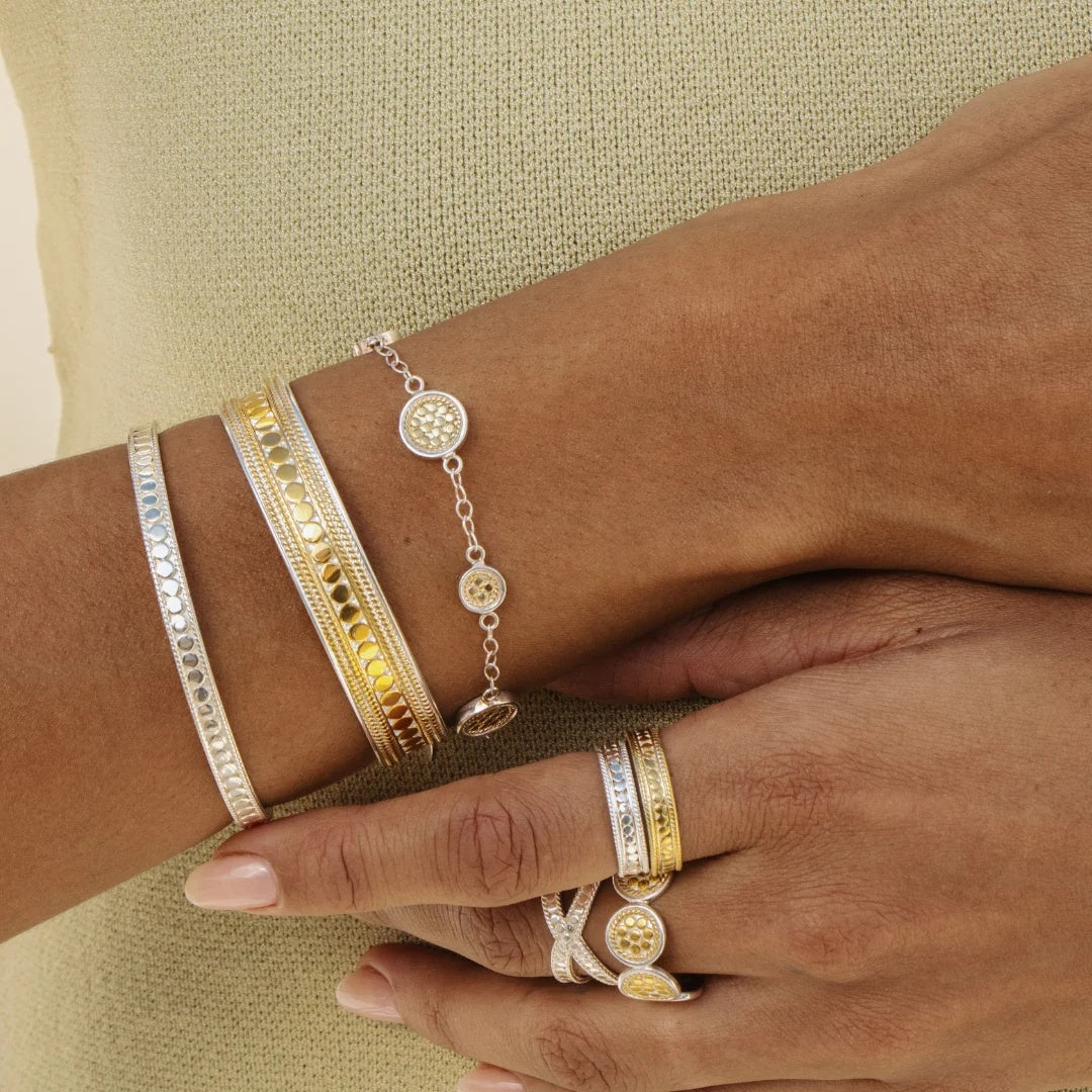 A close-up of a person's hands and wrists wearing multiple delicate bracelets and rings in silver and gold tones, including textured bangles, a chain bracelet with circular charms, and stacked rings with oval motifs.